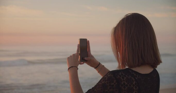 Rear view, side view, young Caucasian woman, taking picture and filming sunset on the beach with her mobile phone. Girl using a smartphone and photographing