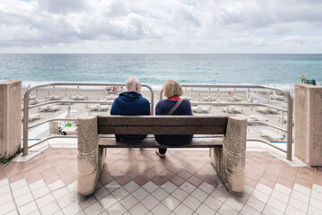 Unrecognizable couple of elderly people, husband and wife, seen from behind, sitting on a bench looking away to the sea and the horizon. Seniors lifestyle.