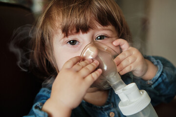 Baby girl in a mask, treatments respiratory tract with a nebulizer at home. Baby sits with a...