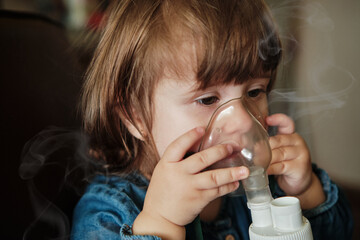 Baby girl in a mask, treatments respiratory tract with a nebulizer at home. Baby sits with a...
