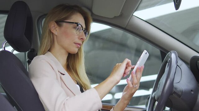 Beautiful blond woman in elegant business suit scrolling through web pages on smartphone sitting in car, doing shopping online, ordering foods and goods sitting at residential building parking lot