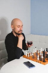 Thoughtful bald man wearing beard playing chess on board near laptop and wineglass. Strategy and competition concept. Side view