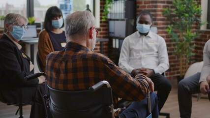 Senior man with disability attending aa group therapy session with people during pandemic. Person with face mask sitting in wheelchair and talking about alcohol addiction at meeting.