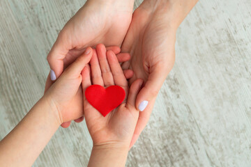 Red heart in child and mother hands on wooden background. Health care and family concept.