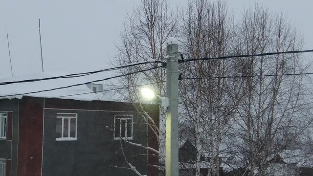 Aerial shot in backward movement from street light when strong snow-storm in evening. Some building and winter trees behind the light