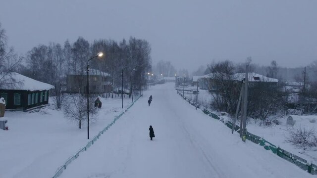 Aerial shot in forward movement over snowed town when strong snow-storm in evening. Road in center of frame. Street lights are on, some people and one car on the street