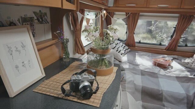 Tracking shot of interior of cozy campervan decorated with fabric flags garland. Flowers in vases, photos and digital camera are on cupboard. Ukulele and pillows are lying on bed