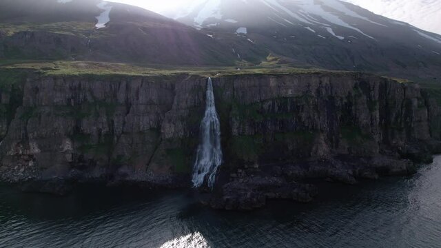 Backwards aerial shot moving away from cliffs with waterfall flowing into ocean. Epic snowy mountains loom over, shot in Iceland. 