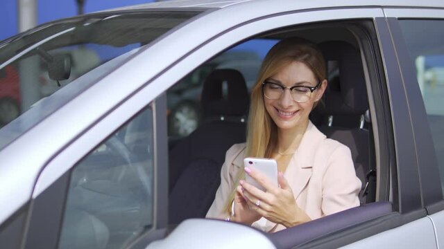 Happy cheerful woman in elegant suit and stylish eyeglasses scrolling web pages on smartphone and smiling, surfing internet sitting in car, birthday girl accepting congratulations online