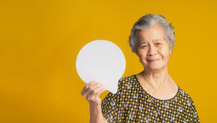 An elderly Asian woman with short gray hair holding a blank speech bubble and looking at the camera with a smile while standing on a yellow background in the studio