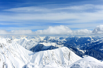 Obraz premium View of snow-covered mountain peaks over which the clouds are floating. In the background is a blue cloudy sky.
