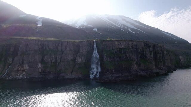 Dramatic revolving aerial shot of waterfall flowing into ocean over cliffs. Shot in Iceland by mountain coastline. Bright sun reflects on water.
