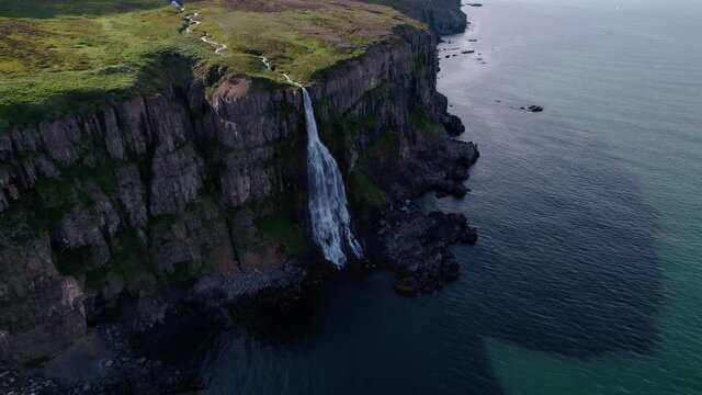 Aerial shot of flowing waterfall on the side of a cliff into ocean, shot in Iceland. Green grass and birds flying below.