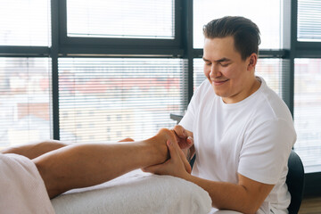 Close-up of cheerful professional male masseur with strong hands massaging feet heels of muscular athlete man after physical sports workout, lying on stomach at massage table in salon by window.