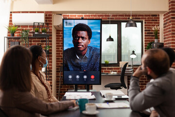 Multi-ethnic group of businesspeople with medical face mask discussing financial strategy with remote collegue. Online videocall meeting conference in startup company office. Business call on monitor