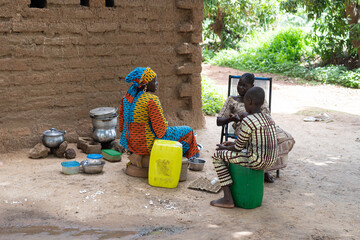 Black African woman in a colourful dress sitting in her outdoor kitchen chatting amicably with...