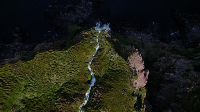 Birds eye aerial view over a waterfall in Iceland, flowing into the ocean. Lush green grass and dark ocean.