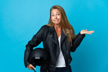 Middle age woman holding a motorcycle helmet isolated on blue background having doubts while raising hands