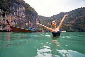 Vacation and happiness. Pretty young woman rising hands up standing on tropical beach with beautiful view in Thailand.