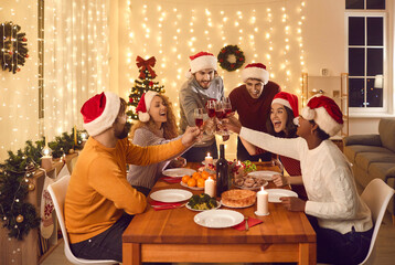 Overjoyed multiethnic people in Santa hats clink glasses dining together at table on Christmas eve. Smiling diverse friends cheers celebrate New Year winter holiday have delicious dinner at home.