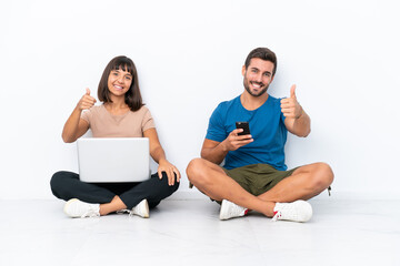 Young couple sitting on the floor holding pc and mobile phone isolated on white background giving a...