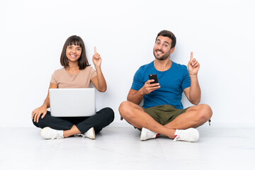 Young couple sitting on the floor holding pc and mobile phone isolated on white background showing and lifting a finger in sign of the best