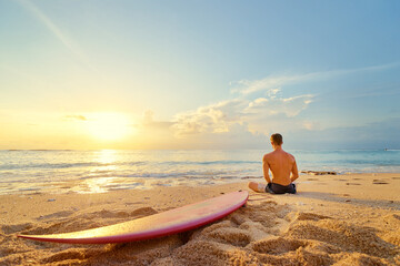 Surfing and meditation. Enjoying sunset. Relaxed young man sitting on lotus position with surf board on the beach.