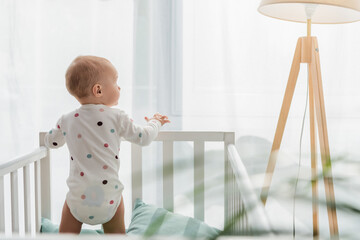 back view of toddler kid in baby romper standing in crib.