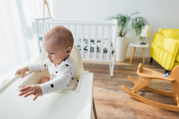 Fototapeta premium toddler boy sitting in baby chair near blurred crib and rocking horse.