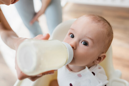 high angle view of toddler boy drinking milk from baby bottle near blurred mom.