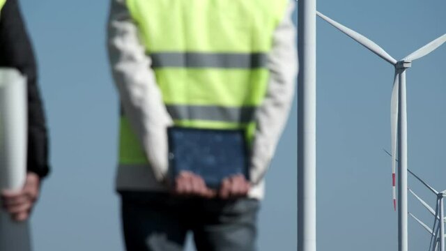 Windmills produce clean energy at offshore station under blue sky. Professional engineer stands holding tablet behind back against rotating turbines