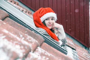 Young woman in a red and white Christmas hat looking out of a skylight window and smiling, propping...