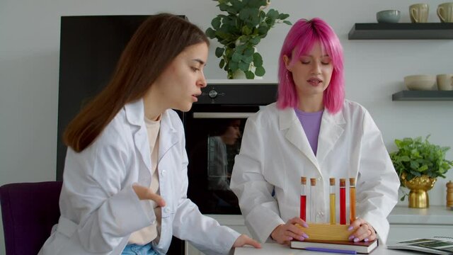 Positive attractive woman with physical disability and beautiful pink haired female researcher wearing lab coats, testing flavor of new perfume from test tube while making scientific research indoors.
