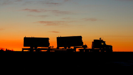 A transportation themed image representing supply chain management and logistics, of a large truck hauling trailers on the highway silhouetted by the sunrise.