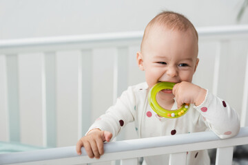 cheerful toddler kid biting rattle ring while standing in crib.