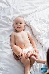 high angle view of toddler boy in diapers lying on bed near blurred mom.