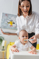 smiling woman holding laptop while feeding baby boy with puree at home.
