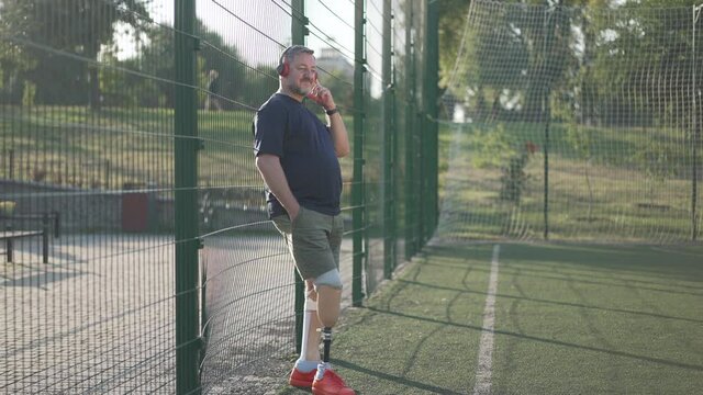 Wide shot portrait happy male Caucasian amputee listening to music in headphones standing at mesh fence on summer sports field. Mature grey-haired man with prosthesis enjoying hobby after training