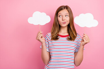 Photo of unsure small brown hairdo girl hold clouds look empty space wear striped t-shirt isolated on pink color background