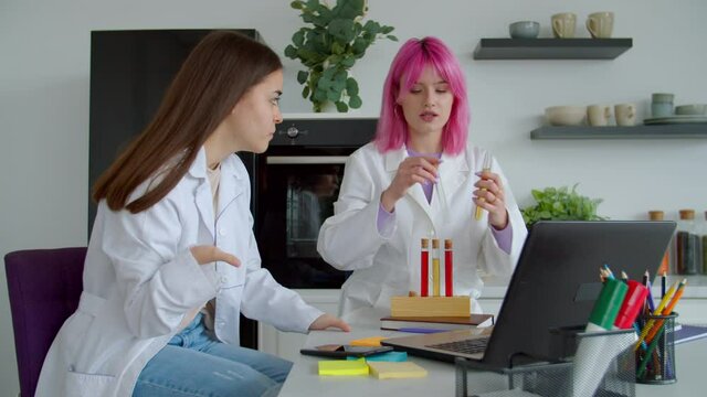 Concentrated pretty pink haired researcher and lovely female student with congenital physical anomaly of hands wearing lab coats, carrying out scientific experiments with test tubes indoors.