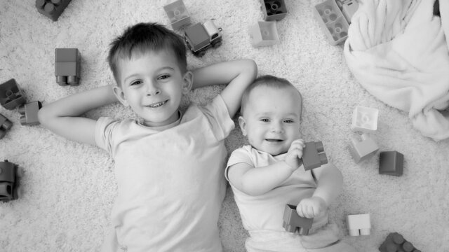 Black and white image of cheerful smiling boy with baby brother surrounded with toys lying on carpet in playroom. Concept of children development, education and creativity at home