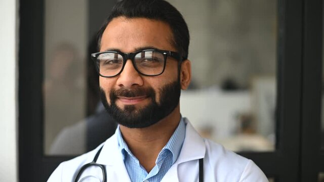Headshot of intelligent professional male Indian doctor, wearing glasses, medical white gown and stethoscope, looking at the camera and smiles, therapist physician standing in the modern clinic