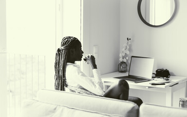 A side view of the silhouette of the stunning female with braids sitting in the wicker chair and drinking an espresso while looking at her laptop screen in the comfortable living room; black and white
