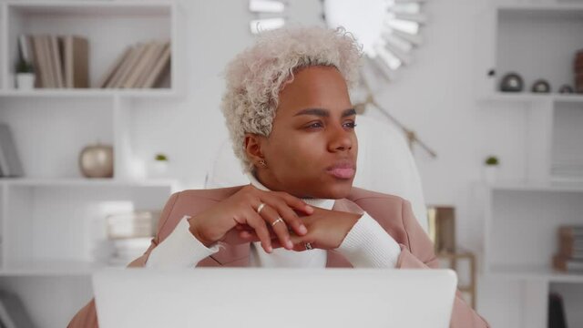 Puzzled African American woman sits with tense face in thought. Attractive brooding female ponders about future, thinks about problems and how to solve them at home office with laptop.