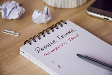 List of passive income generation ideas handwritten in notepad on wooden desk. How to earn extra money concept, selective focus.
