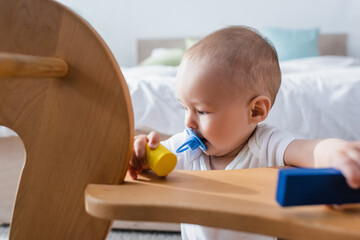 little boy with pacifier holding building blocks near blurred rocking horse.