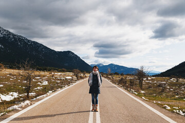 Young smiling woman standing at empty asphalt road in the middle of mountainous area. Highlands of Greece, winter time. Freedom, travel concept