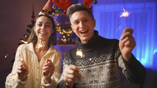 happy family portrait, woman in festive Elf Cap and man in red reindeer antlers headband, looking at camera with sparklers at Christmas or New Year at cozy house. 4k slow motion