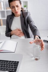 blurred pregnant businesswoman feeling cramp and reaching glass of water on office table.