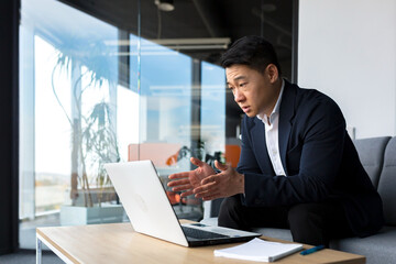 Concentrated male businessman talking to colleagues on video call using laptop sitting in office, asian man working online